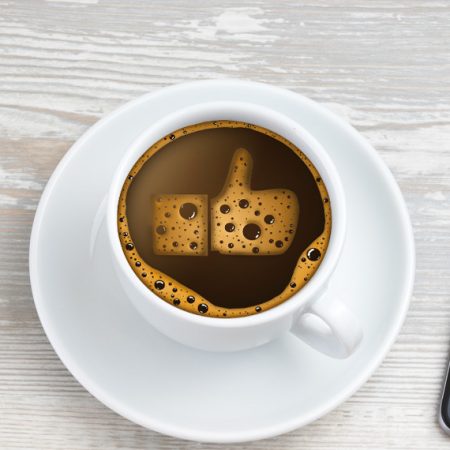 A desktop from above with computer, coffee cup with thumbs up emoji in foam and mobile phone and notepad