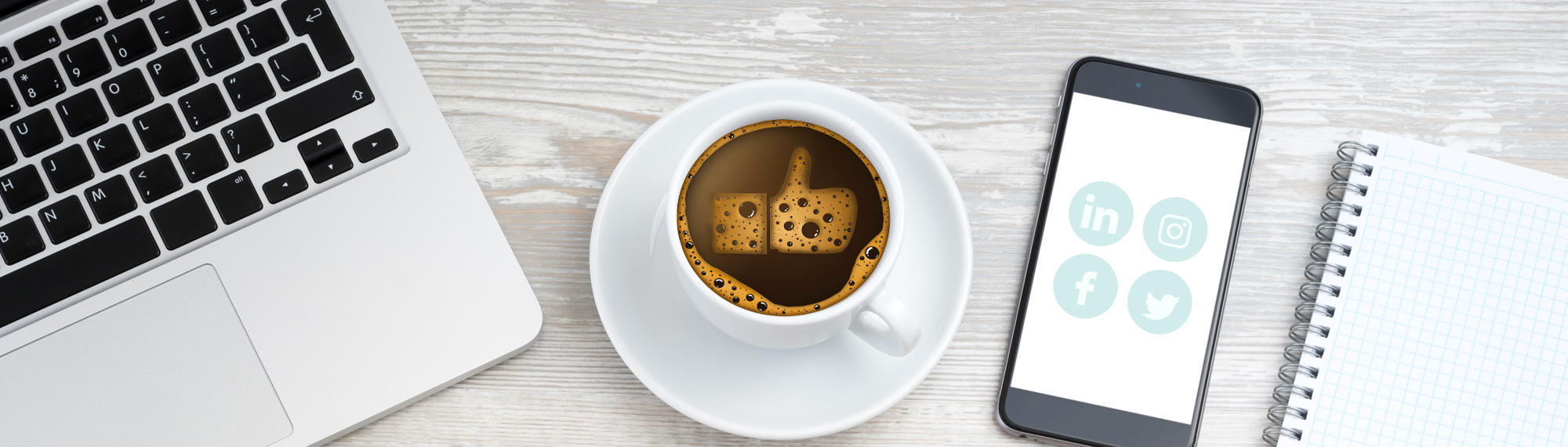 A desktop from above with computer, coffee cup with thumbs up emoji in foam and mobile phone and notepad